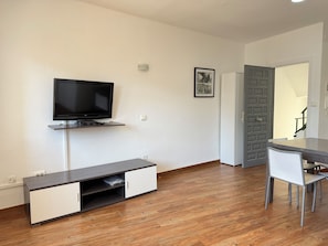 Ein modernes Wohnzimmer mit Flachbildfernseher, einer eingebauten Schrankwand, einem Esstisch mit Stühlen und einer Tür zu einem weiteren Zimmer.