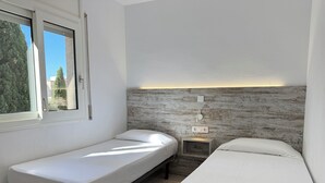Quarto com duas camas de solteiro, uma pequena mesa de cabeceira, uma luminária na parede e uma janela com vista para árvores.