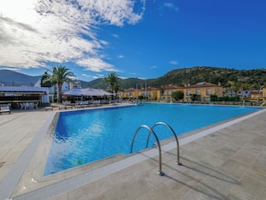 A swimming pool with sun loungers and umbrellas, surrounded by buildings and palm trees.