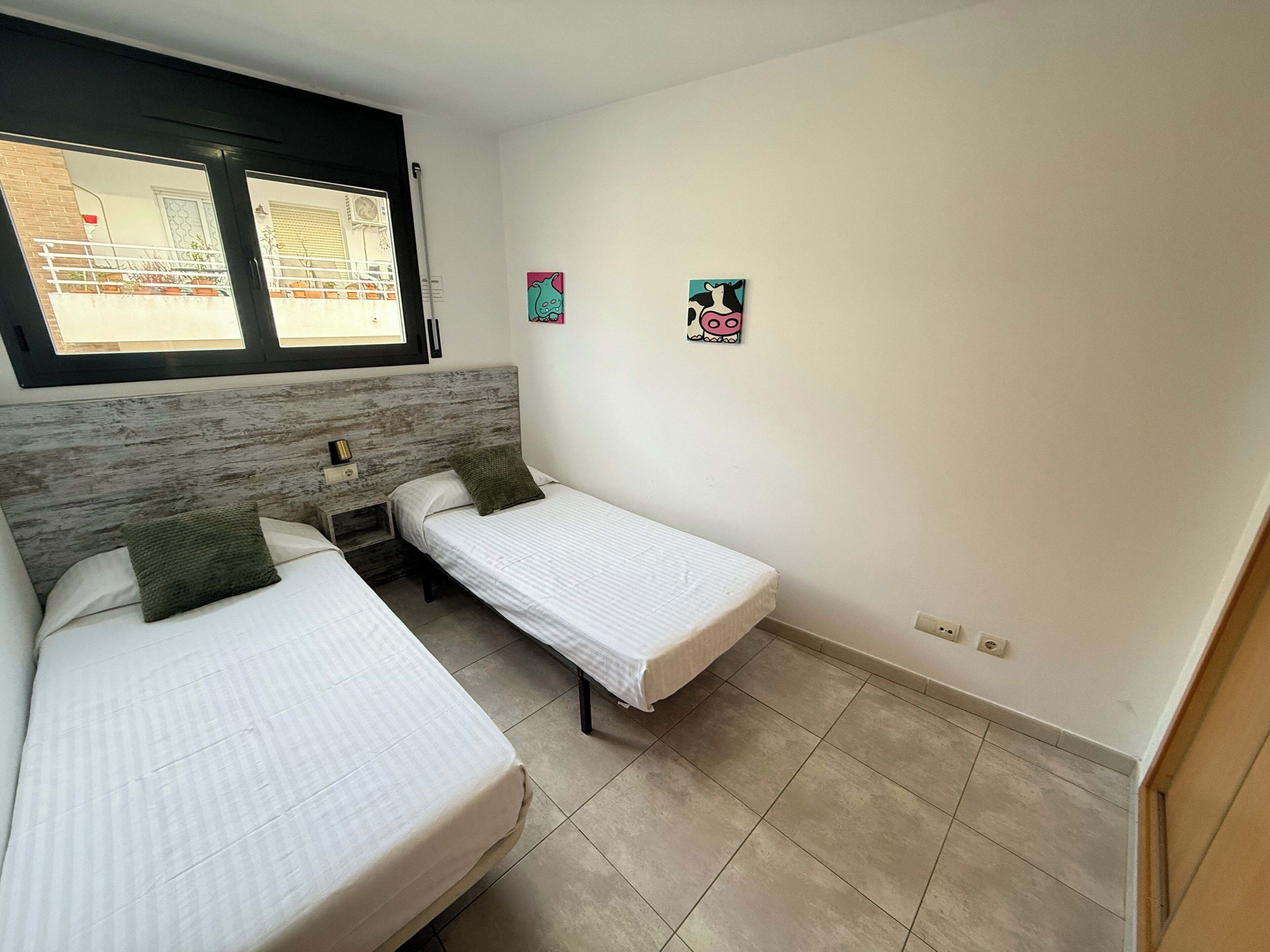 Une chambre avec deux lits simples, une tête de lit en bois, une fenêtre avec des stores et deux éléments de décoration murale.