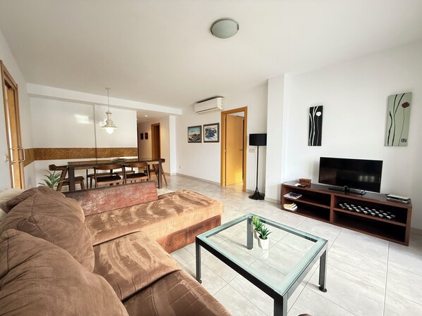 A modern living room with a large sofa, a glass coffee table, a TV stand, and a dining area with a table and chairs. - Agi Riera Ginjolers 3A Apartamento for rent in Roses Girona. (Rosas / Roses)