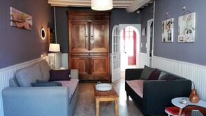 Un salón acogedor con un sofá gris, un sillón oscuro y una mesa de centro de madera. Un mueble de madera con puerta de cristal se encuentra en el centro, y una puerta roja da paso a otra habitación.