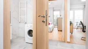 Habitación de hotel moderna con lavadora, puerta de madera y vista hacia un comedor con mesa y sillas.