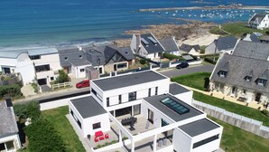 Un quartier résidentiel moderne avec des maisons dotées de toits plats et de grandes fenêtres, une pelouse bien entretenue et une vue sur la mer, où l’on peut apercevoir des bateaux.