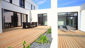 Une terrasse extérieure moderne, dotée d’un plancher en bois, d’un espace salon avec tables et chaises, et d’une porte vitrée donnant sur l’intérieur.