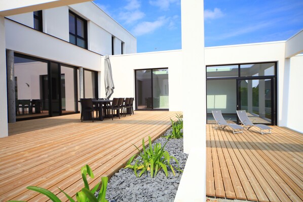A modern outdoor patio with wooden flooring, a seating area with tables and chairs, and a glass door leading to the interior.