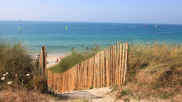 Une plage avec une clôture en bois qui mène au rivage, une eau d’un bleu limpide et des personnes visibles au loin.