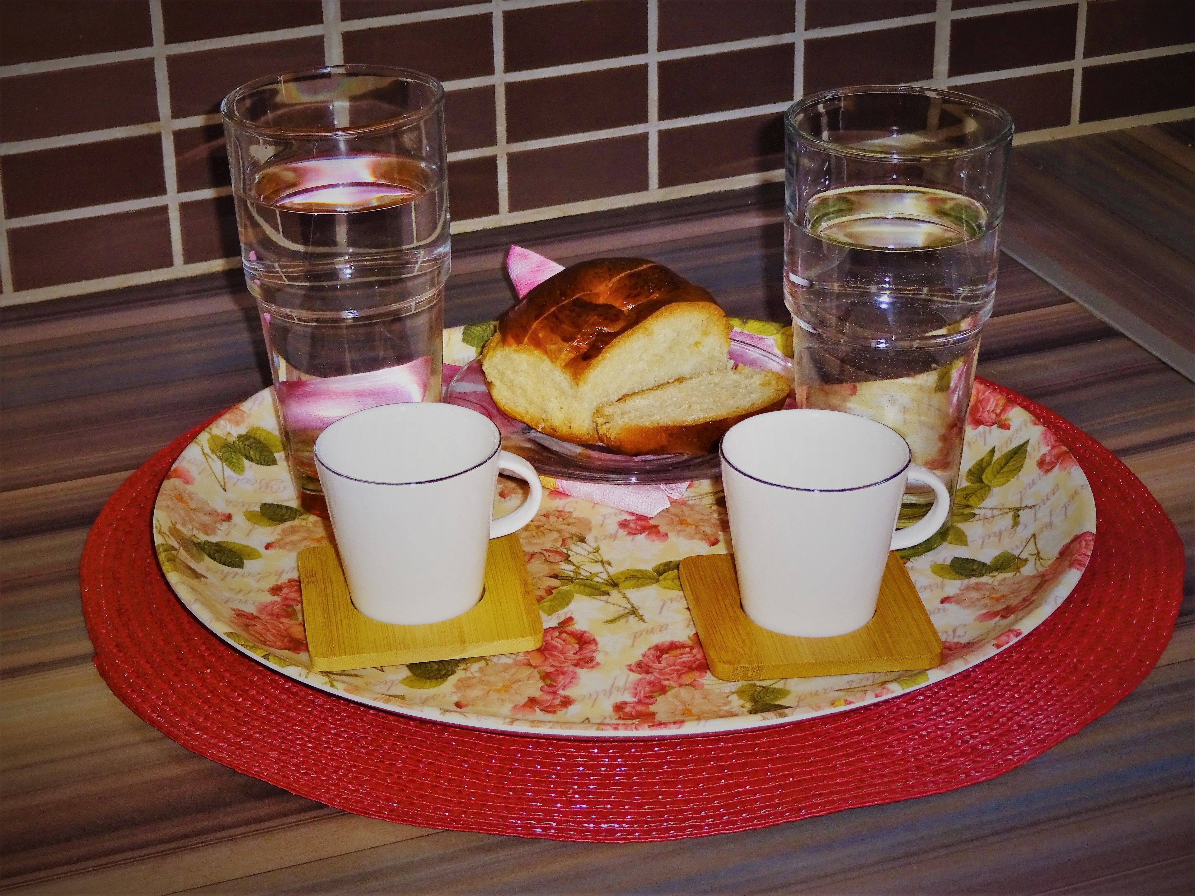 A breakfast tray with two cups, a glass of water, a glass of juice, and a slice of bread on a floral tablecloth.