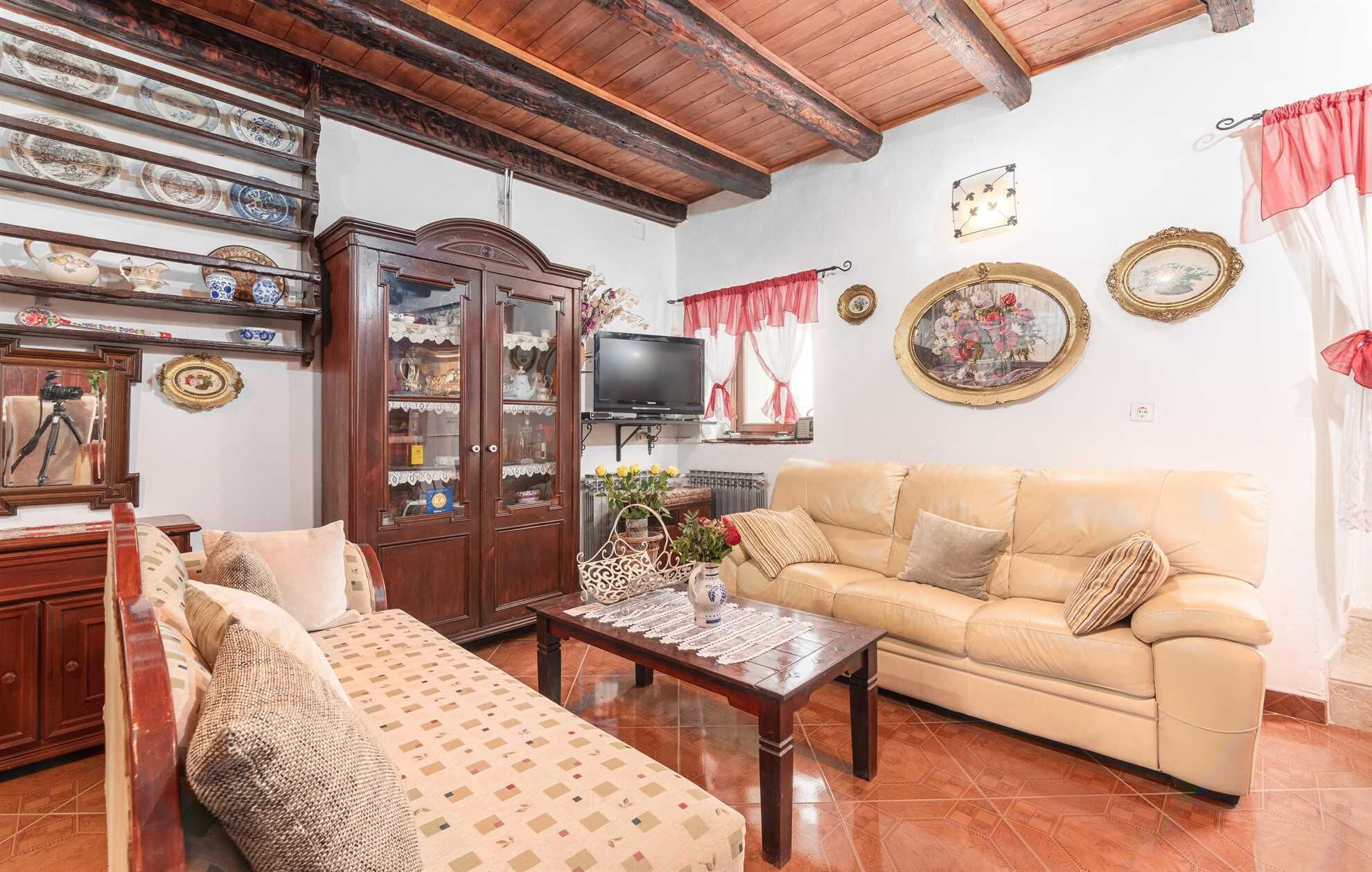 A cozy living room with a sofa, a patterned ottoman, a wooden cabinet with glass doors, and a television.
