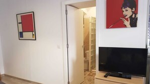 Une pièce avec un téléviseur posé sur un meuble en bois, une porte donnant sur une salle de bain et une œuvre d’art rouge et noire accrochée au mur.