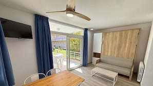 A modern room with a wooden table, white chairs, a sofa, a ceiling fan, and a TV mounted on the wall.