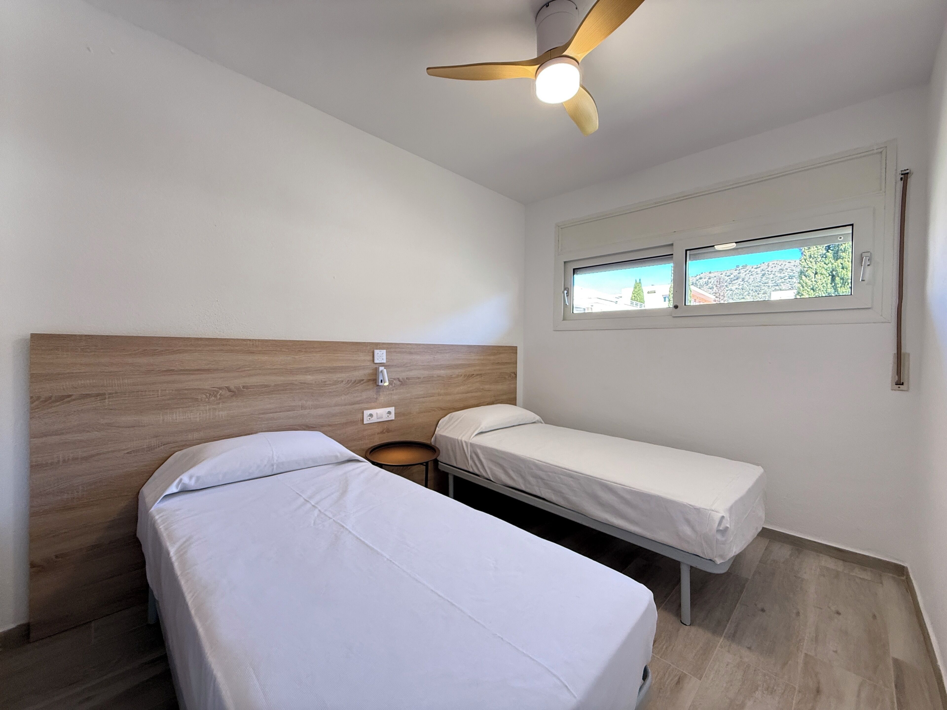 A room with two single beds, a ceiling fan, a window with a view, and a small table.