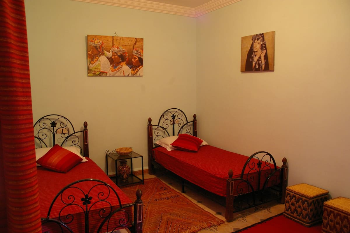 Une chambre avec deux lits simples, une petite table, un tapis décoratif et deux tableaux accrochés au mur.