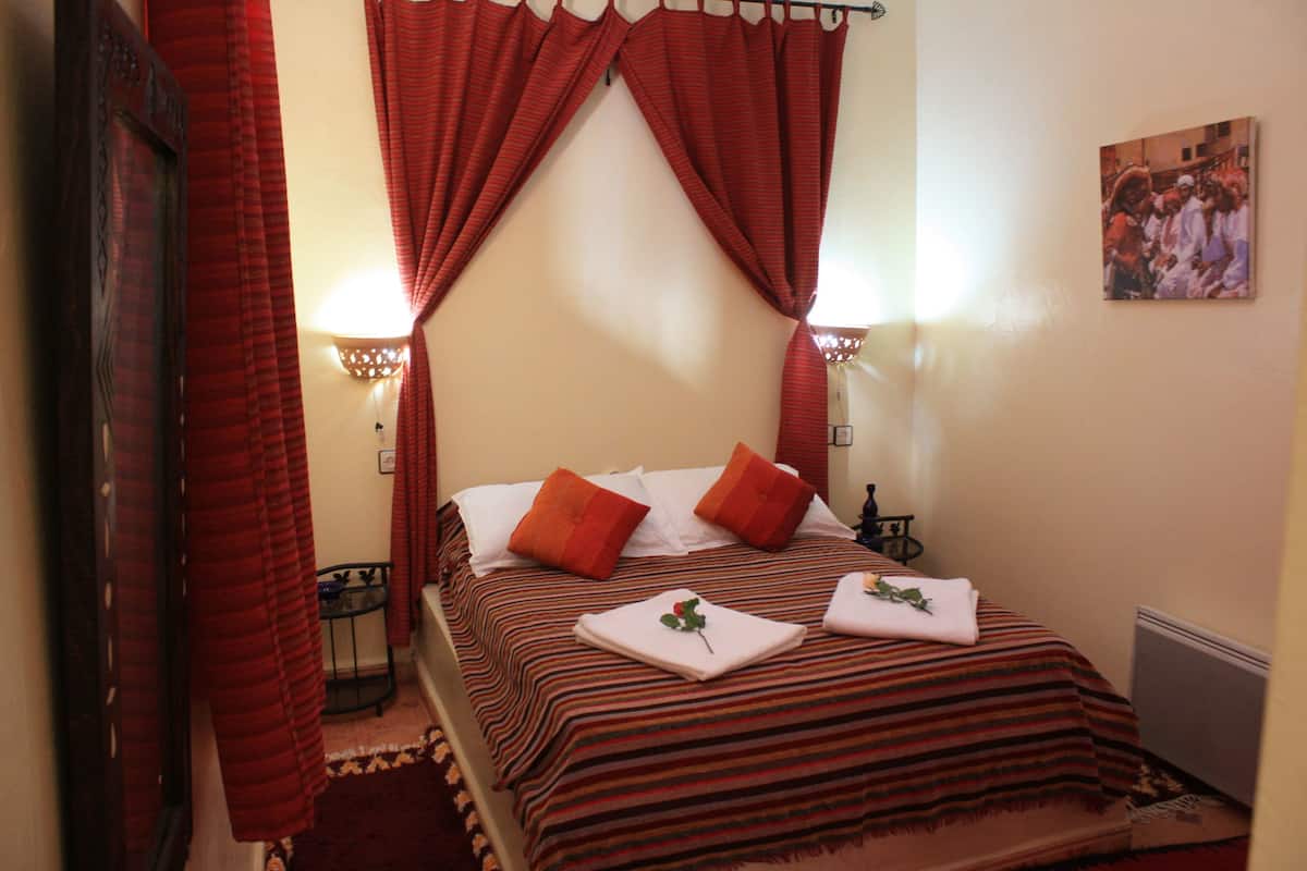 Une chambre d’hôtel avec un lit, des rideaux rouges, deux lampes fixées au mur et un tableau accroché au mur.