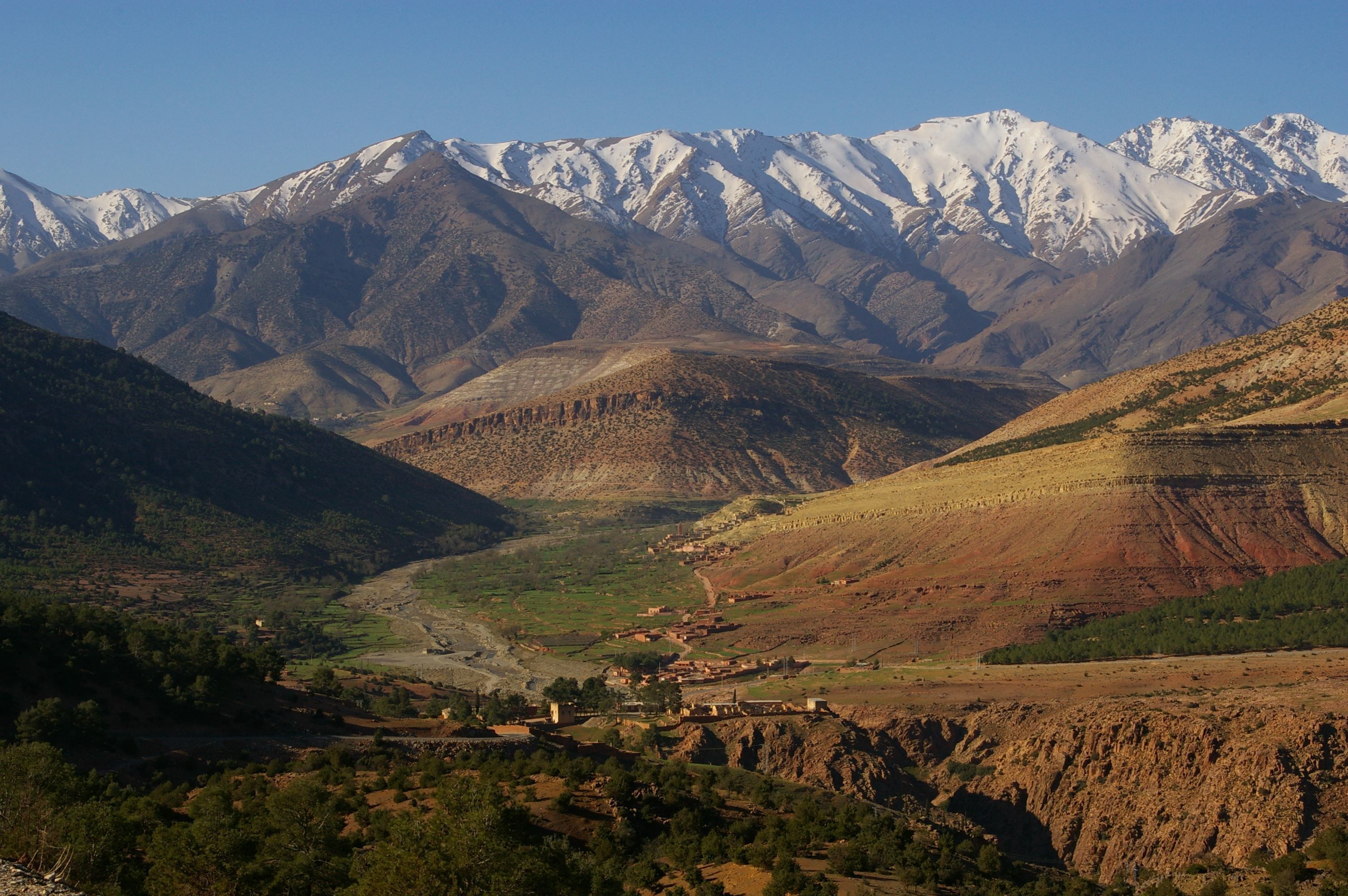 Un paisaje montañoso con picos nevados al fondo, un valle con edificios dispersos y un río que lo atraviesa.
