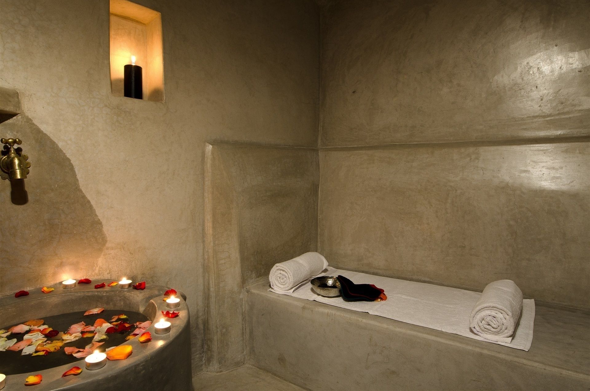 Un baño de paredes de piedra con una bañera, un perchero y un lavamanos con velas y pétalos de flores.