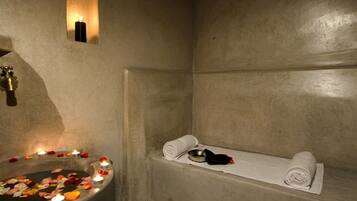Un baño de paredes de piedra con una bañera, un perchero y un lavamanos con velas y pétalos de flores.