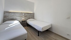 Een kleine, moderne kamer met twee eenpersoonsbedden, een houten hoofdbord, een plafondventilator en een wandlamp.