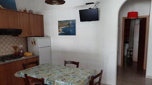 Een keuken met een eettafel, stoelen, een koelkast, een fornuis en een aan de muur bevestigde televisie.