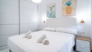 A neatly made bed with white linens and rolled towels, a wooden nightstand with a drawer, and two wall-mounted light fixtures.