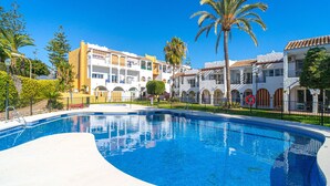 A swimming pool surrounded by multi-story buildings with balconies, palm trees, and a clear blue sky.