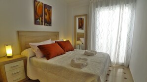 A neatly made bed with white and red pillows, a bedside table with a lamp, and artwork on the wall. - 2 Bedrooms | Trancos | CG R1315 (Nerja)
