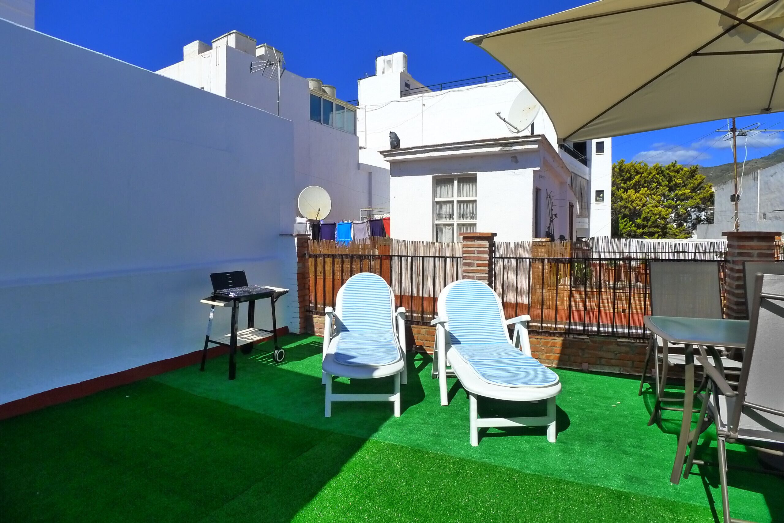 Un patio exterior con césped artificial verde, dos sillones de descanso, una mesita y un ordenador portátil sobre un atril.