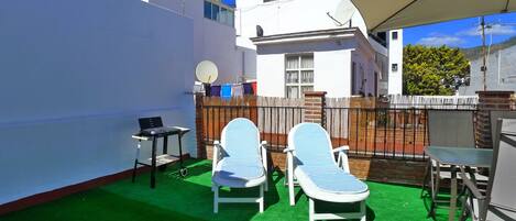Un patio exterior con césped artificial verde, dos sillones de descanso, una mesita y un ordenador portátil sobre un atril.