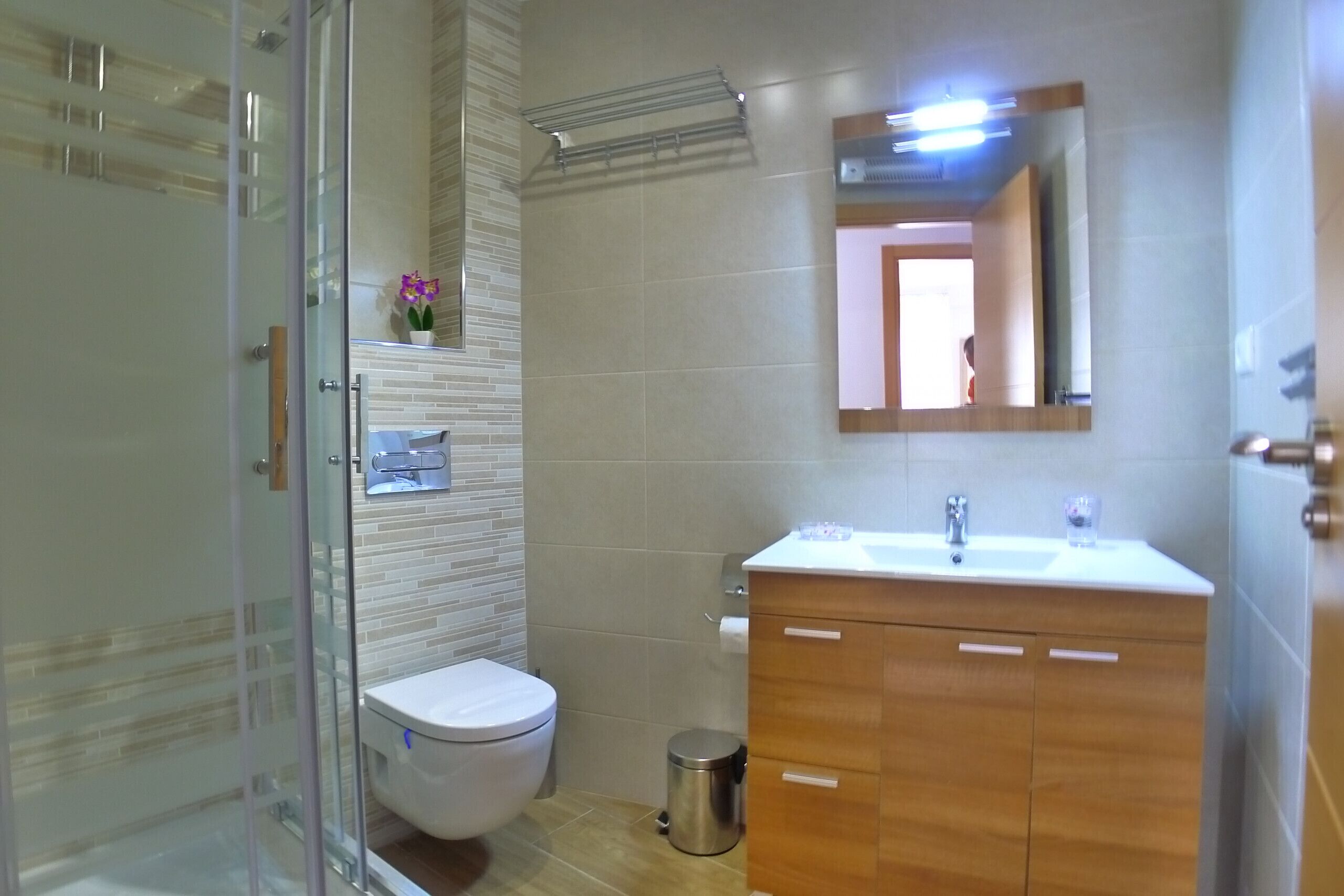 A bathroom with a glass shower enclosure, a toilet, a sink with a mirror above it, and a trash can.