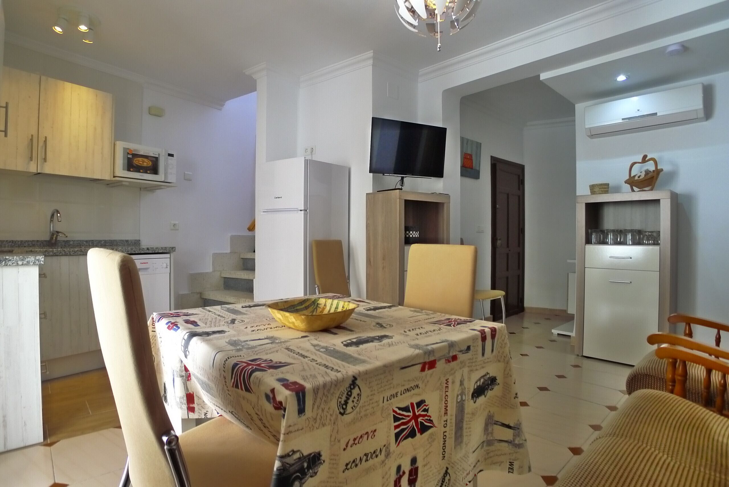 Un espacio habitable compacto con comedor que incluye una mesa con mantel de temática de viajes, una cocina con electrodomésticos modernos y un televisor montado en un mueble de madera.