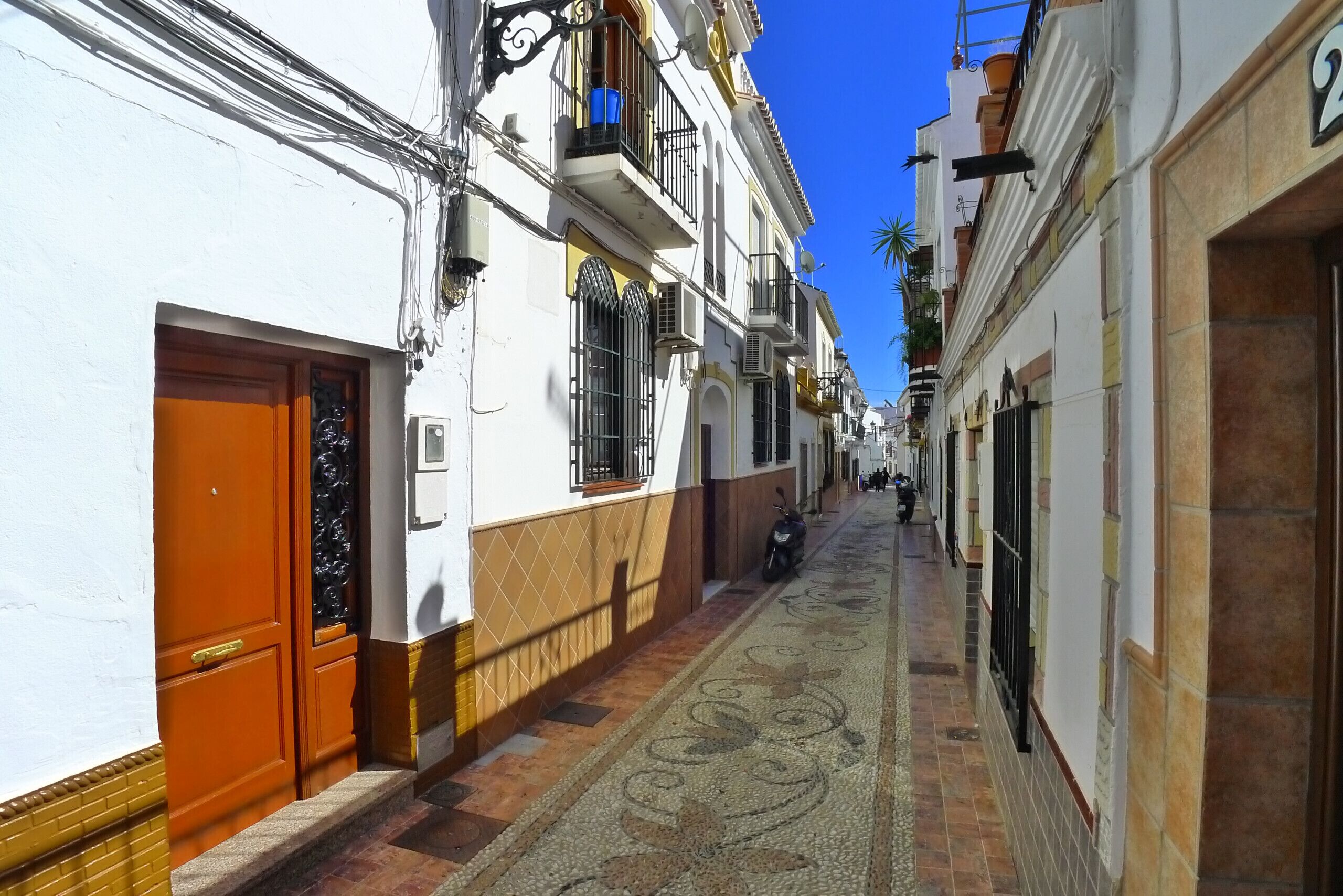 A narrow street with white buildings, a patterned pavement, and a motorcycle parked on the side.
