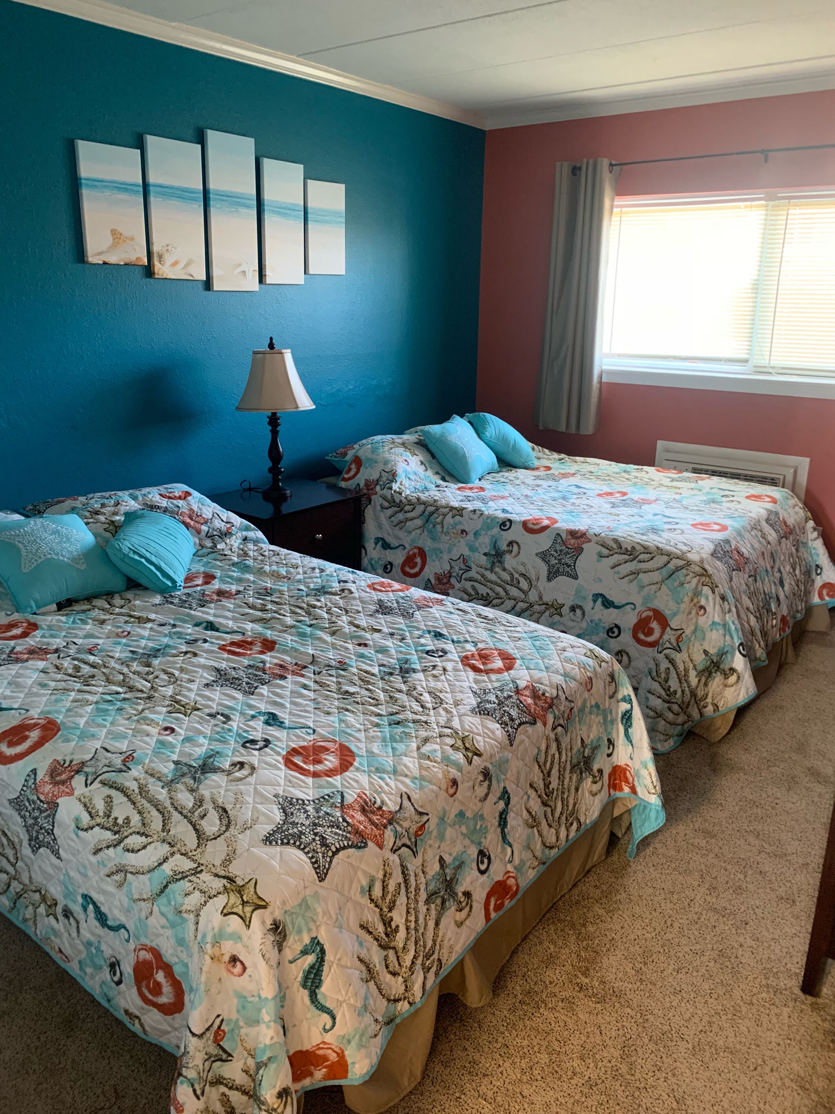 Une chambre avec deux lits, chacun recouvert d’une couvre-lit à motifs floraux, avec une lampe et une fenêtre équipée de stores.