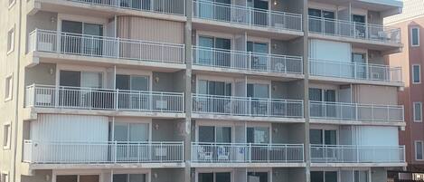 A multi-story residential building with balconies, a sandy ground, and a clear sky.
