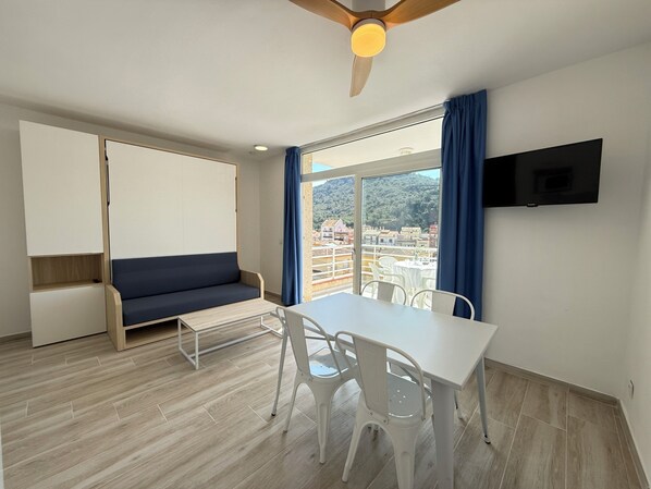 A room with a sofa, a table, chairs, a ceiling fan, a TV, and a large window with a view. - Agi Rescator 215 Apartamento for rent in Roses Girona. (Rosas / Roses)
