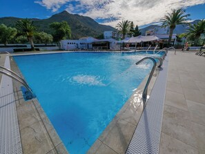 Een zwembad met helderblauw water, omringd door palmbomen en een berg op de achtergrond.