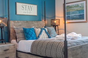 Un lit bien fait, avec une literie à motifs et des coussins décoratifs, une table de chevet avec une lampe, et un mur orné d’un tableau et d’une affiche encadrée.