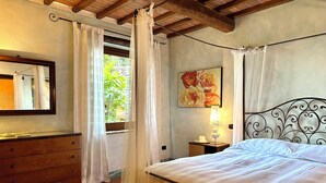 A bedroom with a bed, a dresser, a nightstand with a lamp, a painting, and a window with curtains.