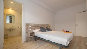 Un dormitorio con cama, mesita de noche y tocador. Hay un baño con lavabo y espejo. La habitación tiene suelo de madera y paredes blancas.