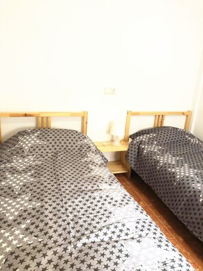 Ein Zimmer mit zwei Einzelbetten, Sternenmuster auf der Bettwäsche, hölzernen Kopfteilen, einem kleinen Nachttisch mit Lampe und einer weißen Wand.