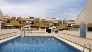 A swimming pool with a geometric tile pattern, surrounded by a fence and a row of yellow buildings with balconies. - BUNGALOW WITH TERRACE 50M2,500M TO THE BEACH (Los Arenales del Sol)