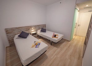 A small, clean room with two beds, a wooden headboard, a nightstand with a lamp, and a door with a green frame.