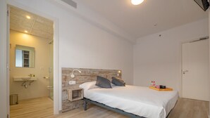 Un dormitorio con cama, mesita de noche y tocador. Hay un baño con lavabo y espejo. La habitación tiene suelo de madera y paredes blancas.