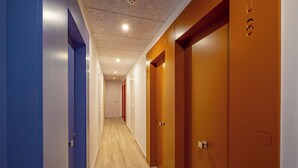 Un pasillo con suelo de madera, puertas azules y anaranjadas, y un techo con iluminación empotrada.