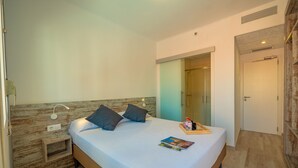 Un dormitorio moderno con cabecera de madera, una cama individual con dos almohadas, una mesita de noche, una lámpara de noche, una revista sobre la cama y un baño con puerta de cristal.