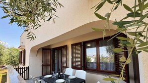 Un área de comedor al aire libre con una mesa preparada para cuatro personas, rodeada de sillas blancas, frente a un edificio con ventanas grandes y contraventanas.