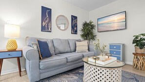 A living room with a grey sofa, a round glass coffee table, a blue and white patterned rug, a wooden side table, a lamp, a mirror, and wall art. - Charmingly decorated beachside condo (Gulf Shores)