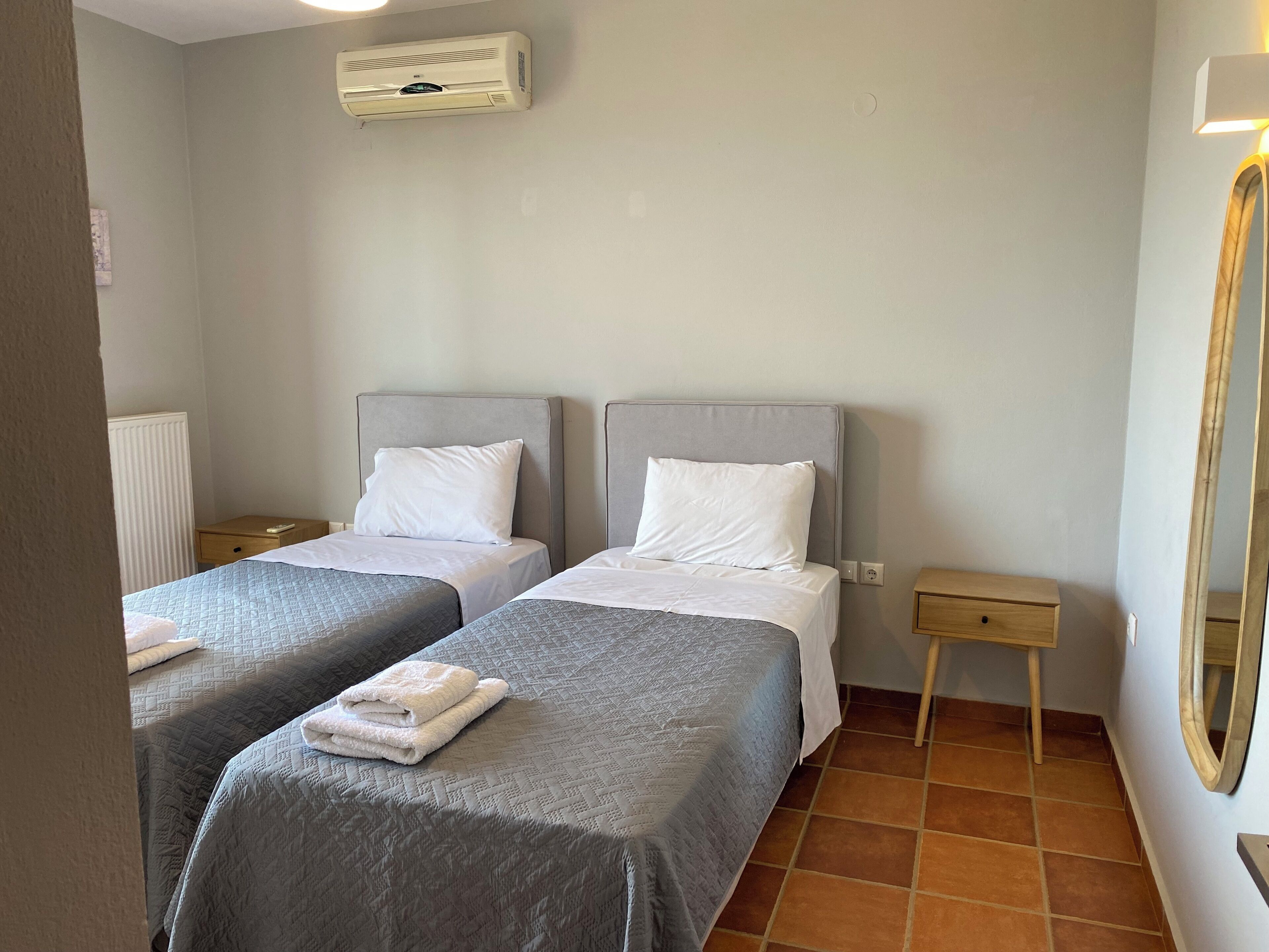 Une chambre d’hôtel avec deux lits simples, une petite table de chevet, une commode, un climatiseur fixé au mur et un miroir.