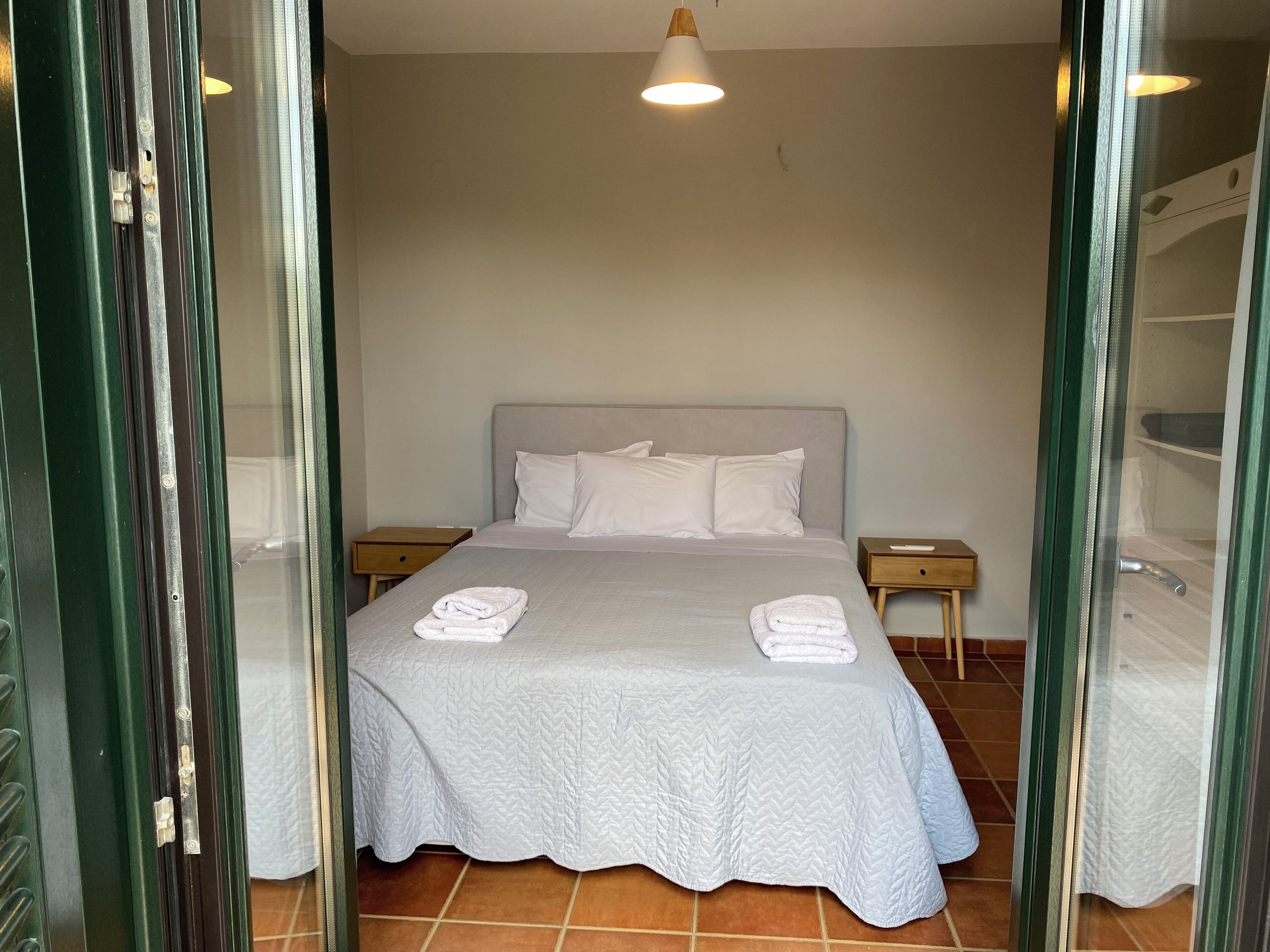Uma cama bem arrumada com lençóis e almofadas brancas, uma mesa de cabeceira com controle remoto e um lustre.