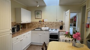 A compact kitchen with white cabinets, a stove, oven, and a sink. There is a table with a vase of flowers and a chair. - Strathan House, Attadale Holiday Cottages, Strathcarron, Ross-shire (Strathcarron, Wester Ross)