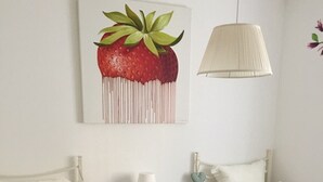A hotel room with two single beds, each with a green bedspread and floral pillows. There is a small bedside table with a lamp, a heart-shaped decoration, and a folded towel on each bed. A large painting of a strawberry hangs on the wall above the beds. - DREAM HOLIDAY ON LAKE GARDA (Lazise)